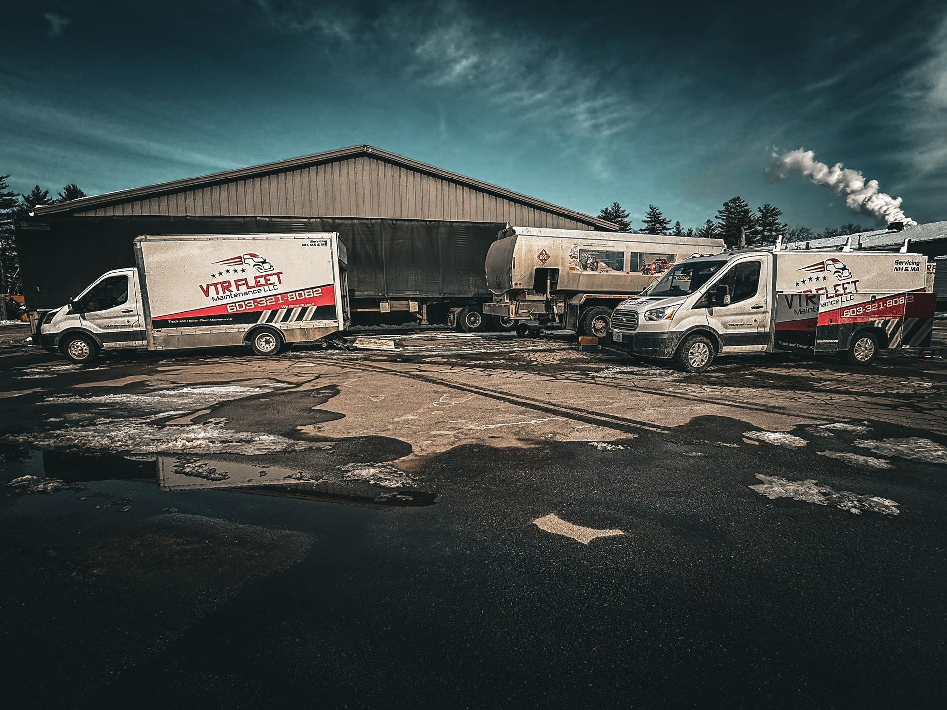 Two white service vans with logos parked near a building and tanker. Smoke rises from a stack.