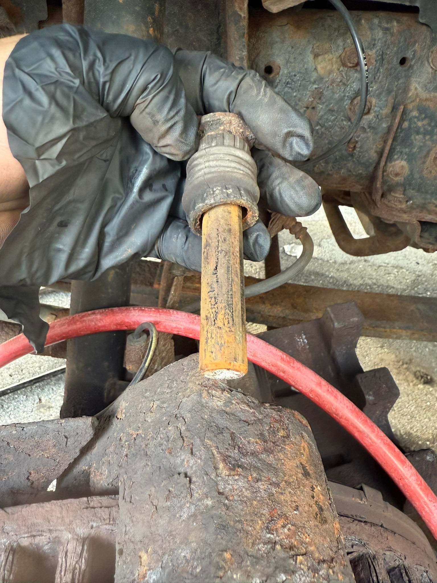 Gloved hand holding a rusty metal part near a vehicle's undercarriage; red hose visible.