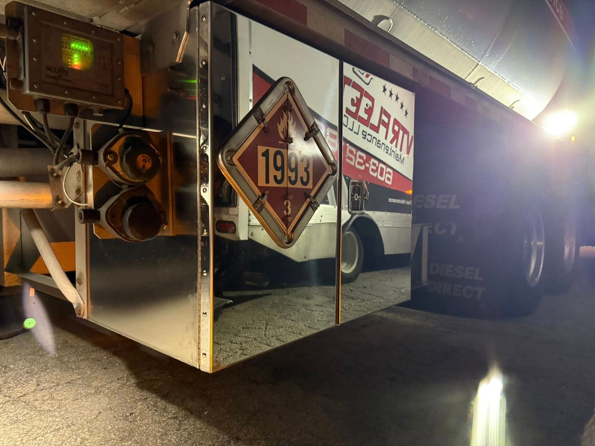 Side of tanker truck with a reflective surface showing a 1993 hazard placard and its reflection of another vehicle.