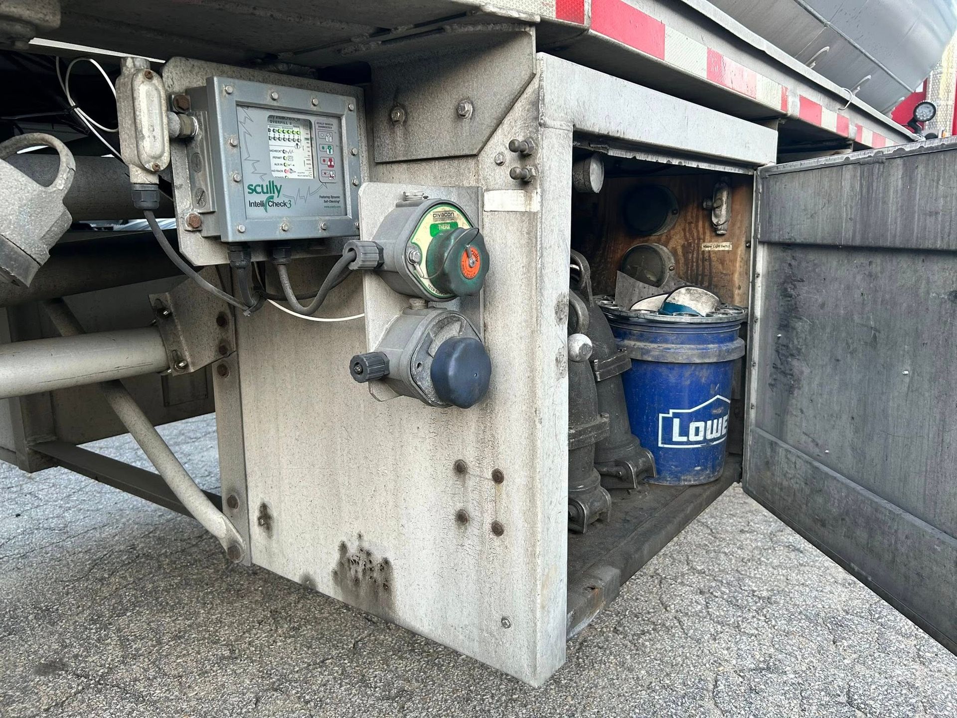 Control panel and access door on a tanker truck, with gauges, wiring, and a blue bucket.