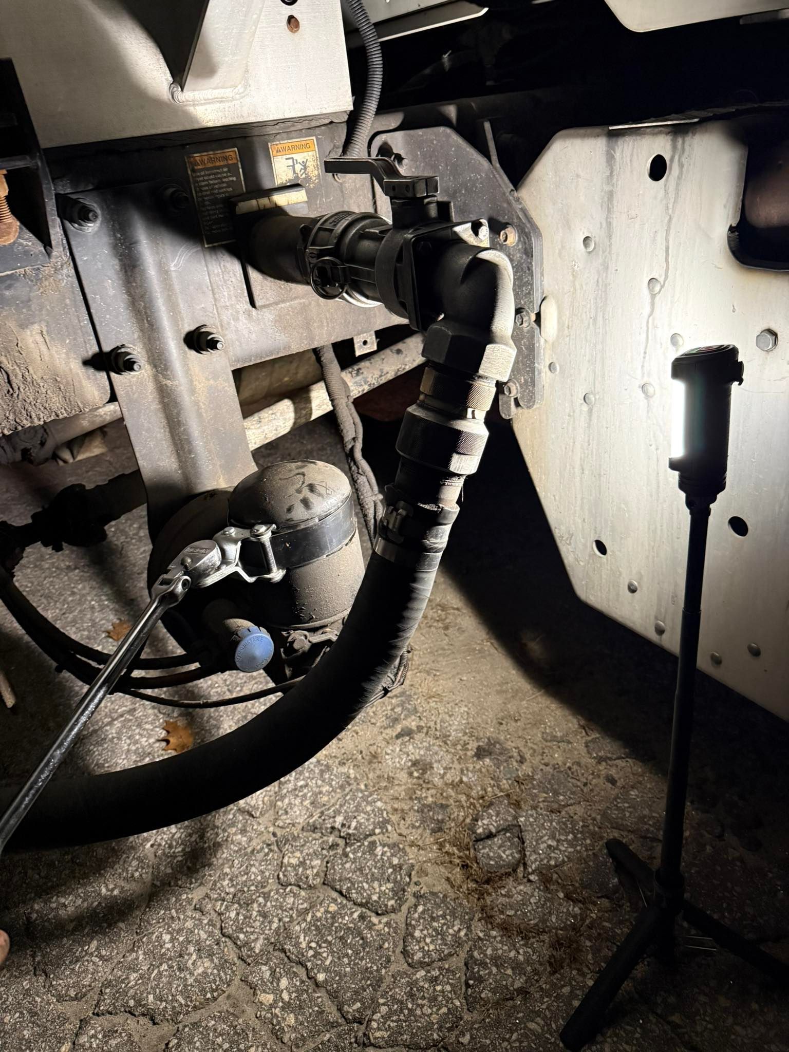 Underside of a vehicle. A black hose is connected to metal pipes, illuminated by a work light.