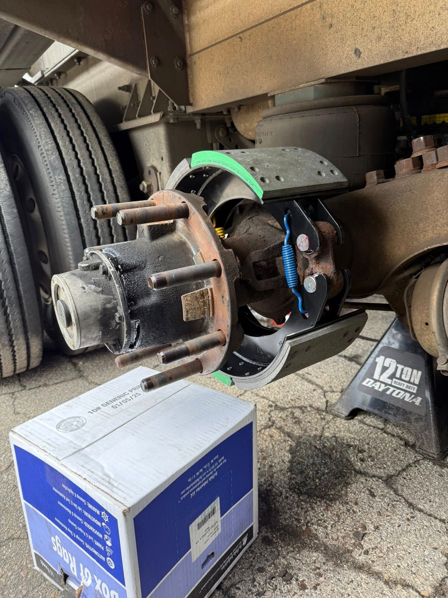 Truck wheel hub being serviced with new brake pads and hardware.  Box of parts on ground.