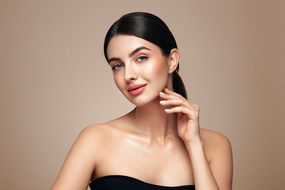 Woman with dark hair in a ponytail, smiling, touching her cheek, against a beige backdrop.