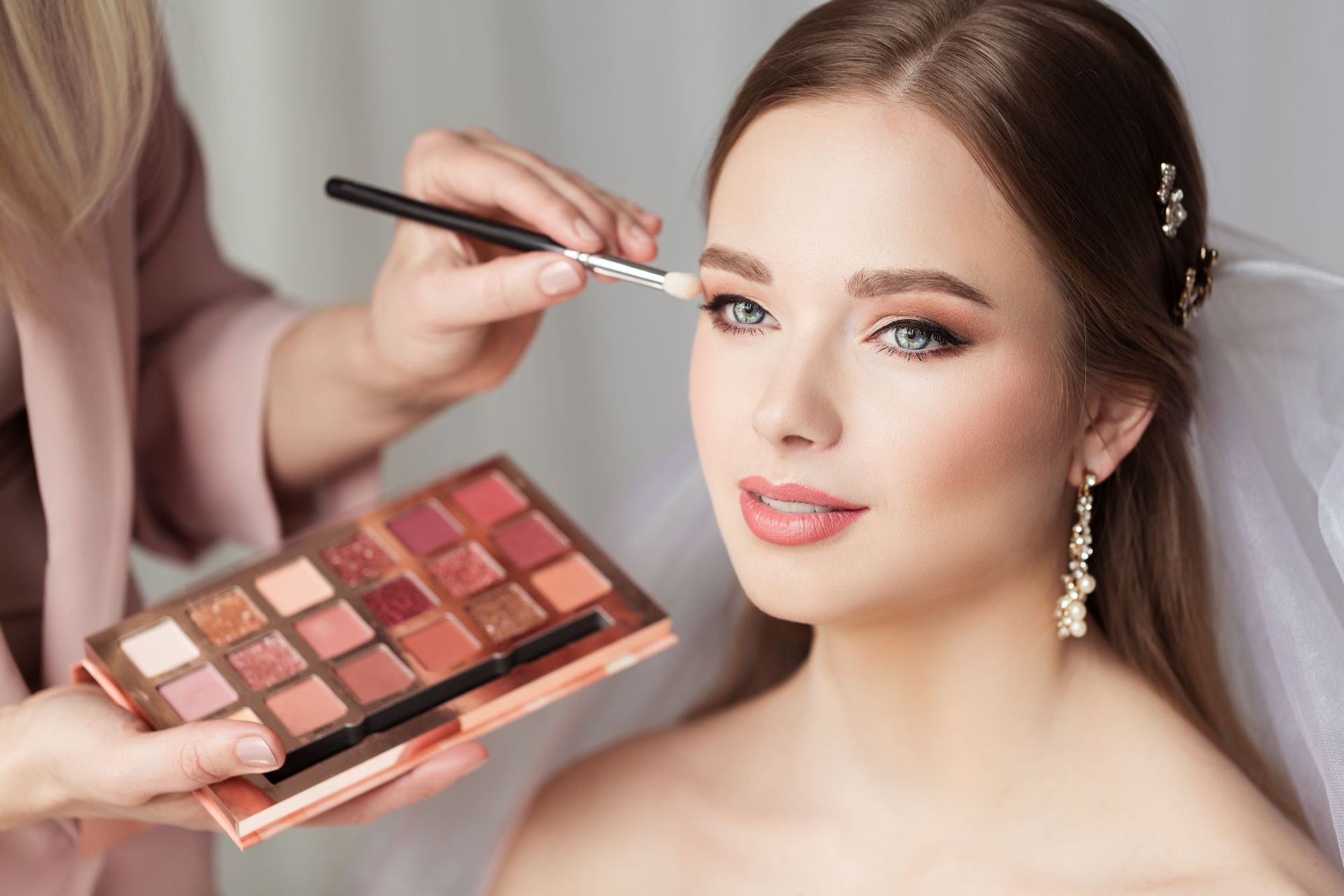 Makeup artist applying eyeshadow on a woman wearing a veil; neutral tones, soft focus.