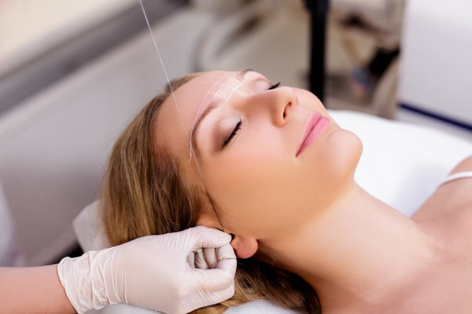 Woman getting eyebrow threading at a salon; technician with gloves uses thread on the brow; close-up.