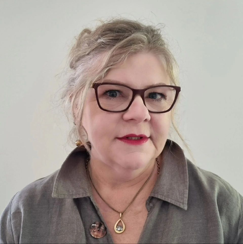 Woman with glasses and dark red lipstick wearing a gray shirt and a necklace.