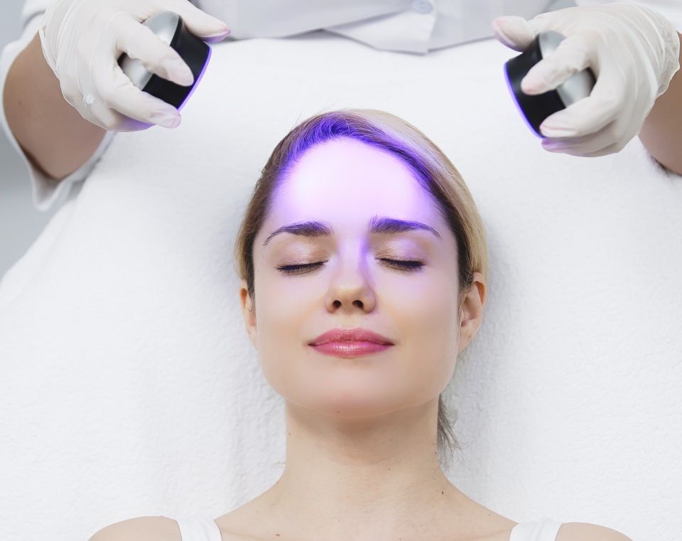 Woman undergoing facial light therapy with devices held by gloved hands.