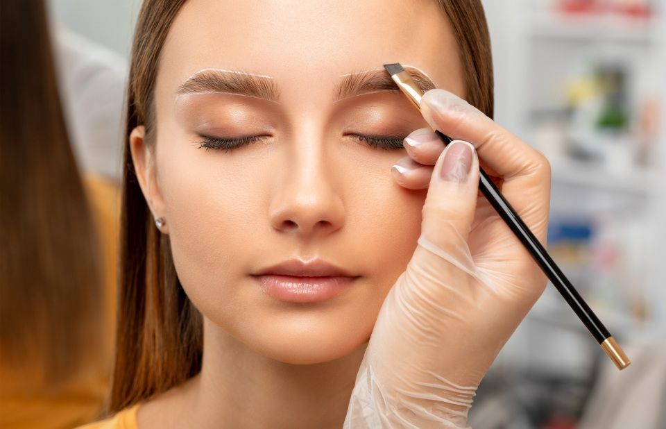 Person's eyebrow being shaped with a brush, eyes closed. Person's hand wears a glove. Bright, indoor setting.