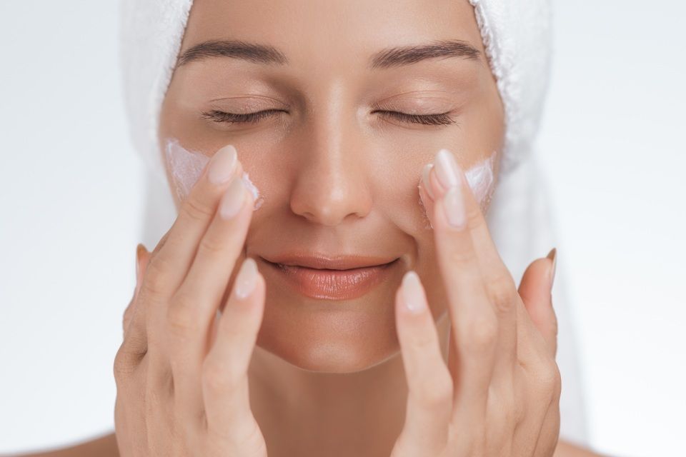 Woman with towel on head applying cream to her face, eyes closed.