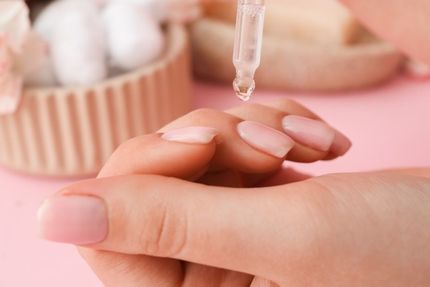 Hand applying nail oil with a dropper. Pink nails, pink background, spa setting.