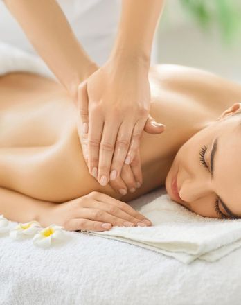 Person receiving shoulder massage at a spa; hands kneading shoulder on white towel, flowers.