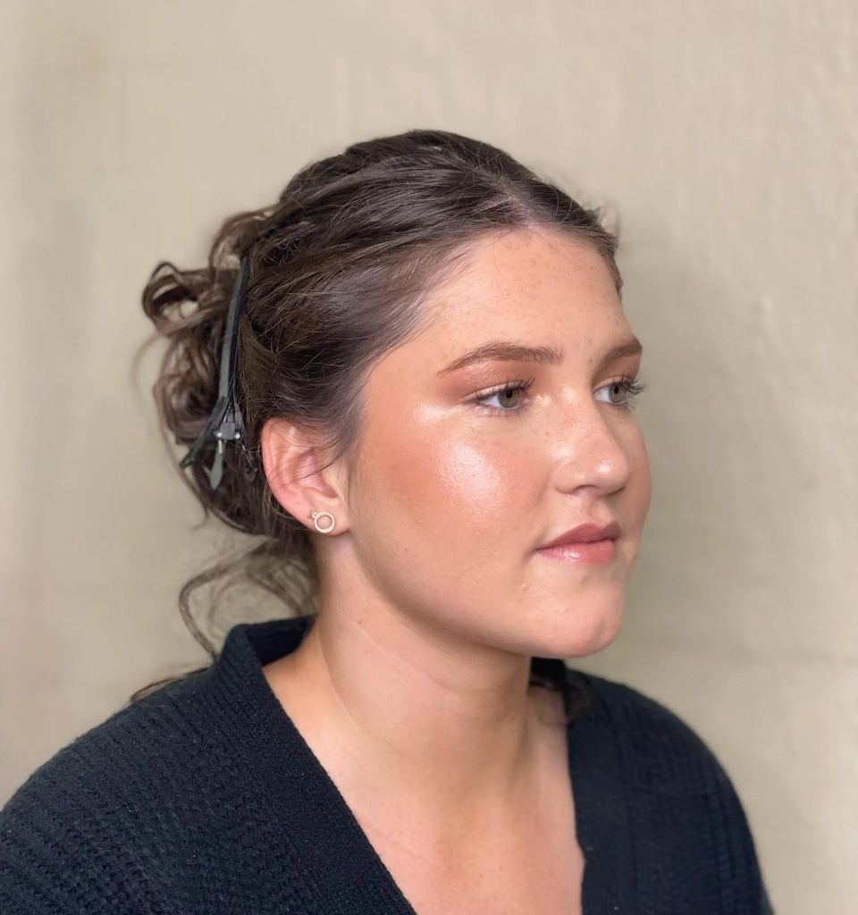 Woman with updo hairstyle and highlighted makeup. She wears a black sweater, side profile.