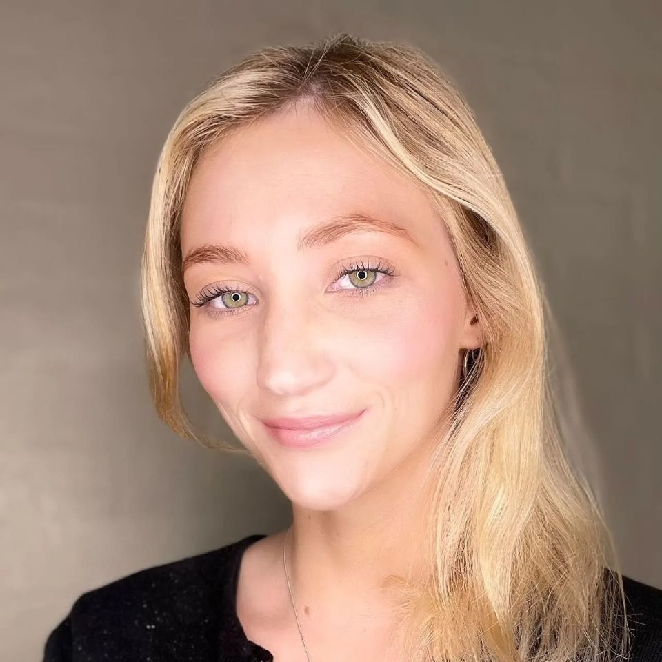 Blonde woman with green eyes smiles; she wears a black shirt, set against a grey background.