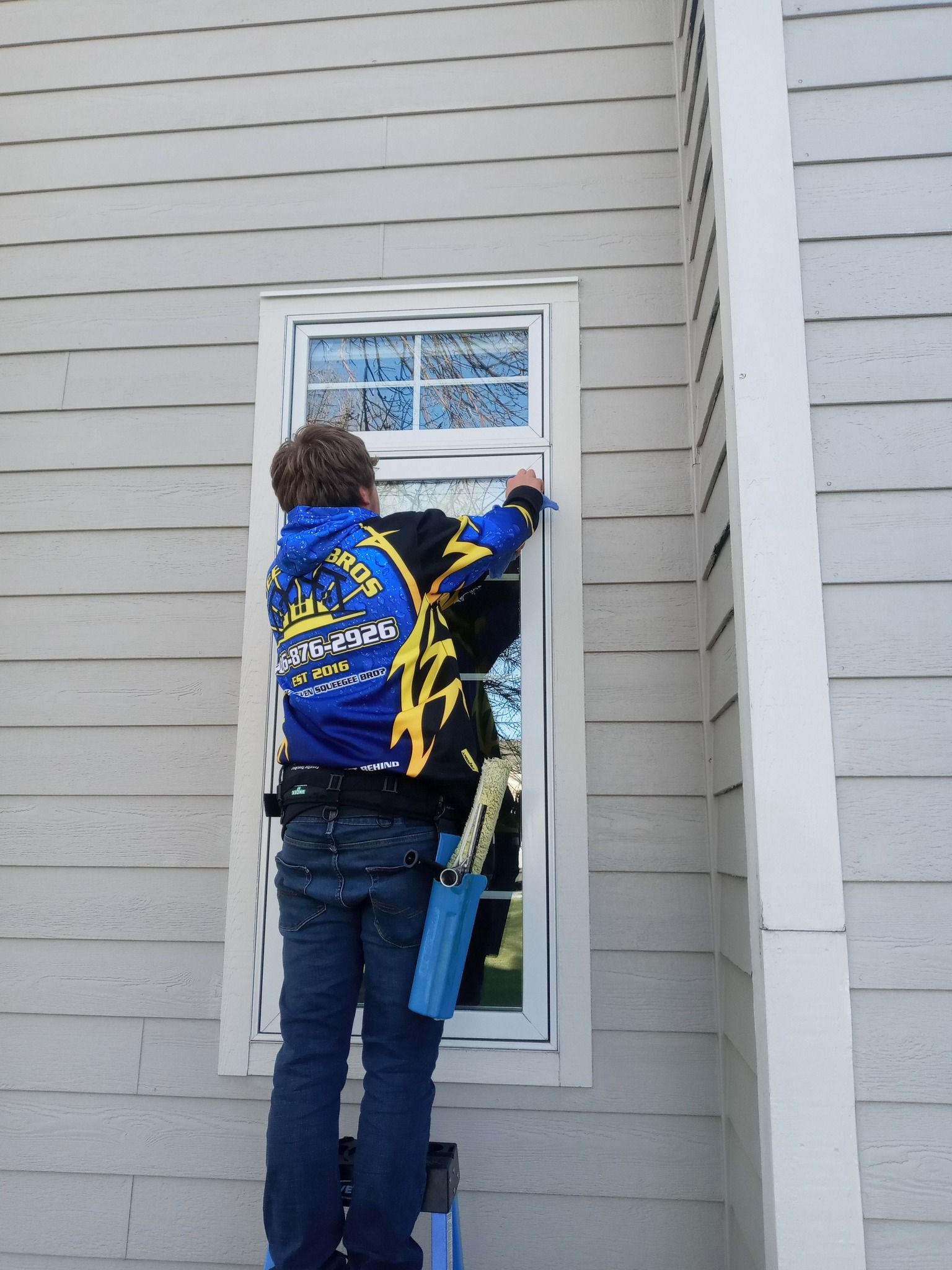 A man standing on a ladder cleaning a window with a sweatshirt that says ' ubc ' on the back