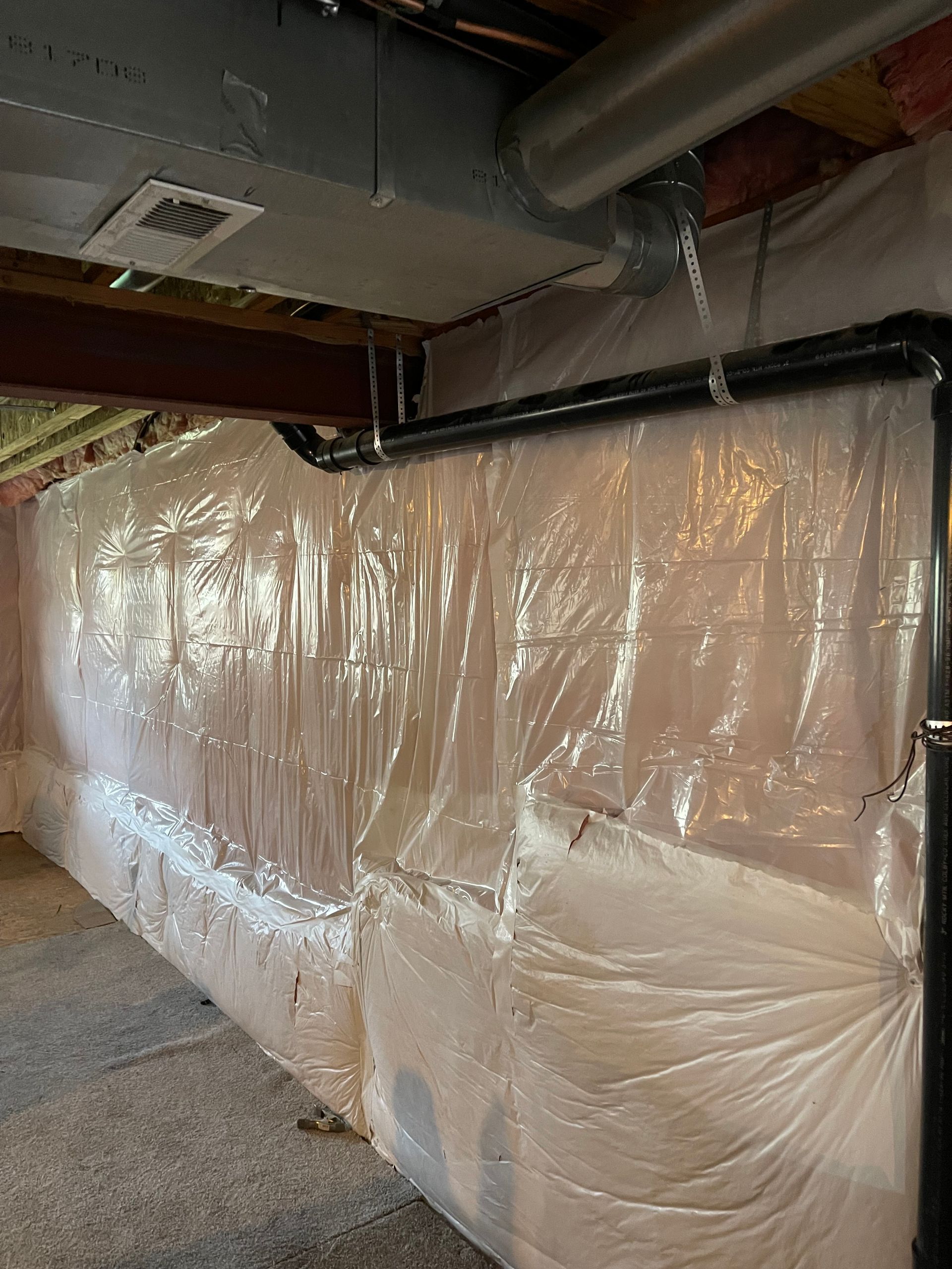 Basement wall lined with clear plastic sheeting beneath exposed pipes and ducts