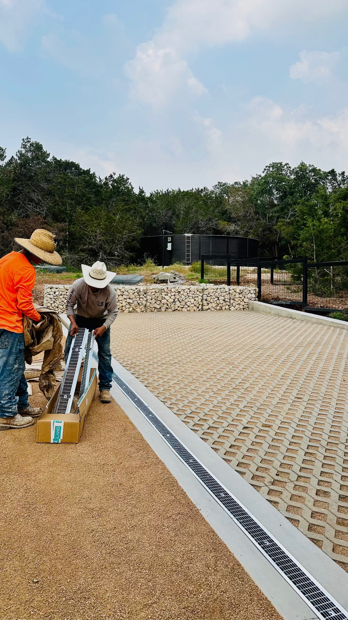 Two workers spreading gravel on a landscaped path beside a white-edged bed and trees