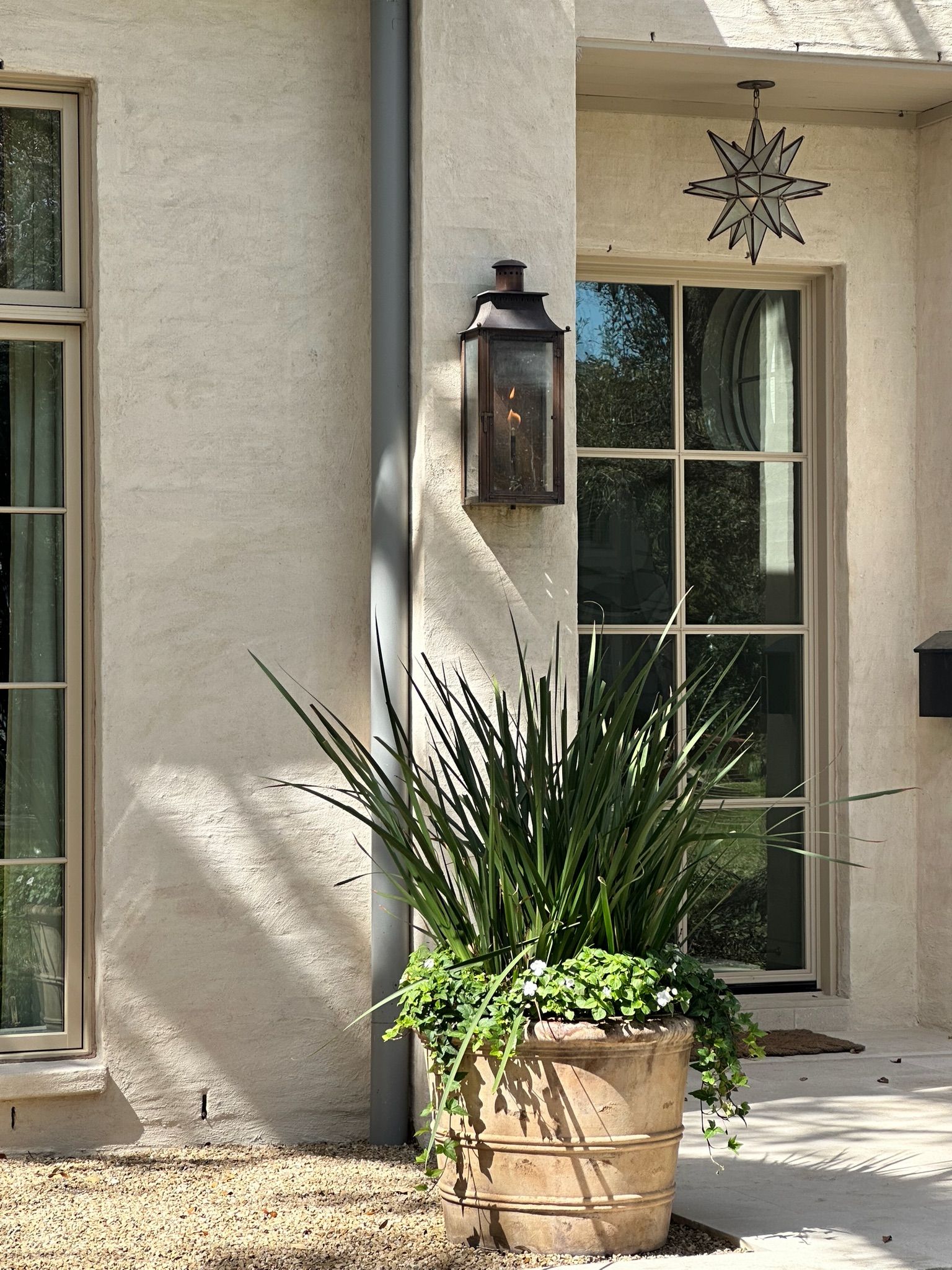 Front porch with a potted spiky plant, wall lantern, and window beside a beige stucco wall