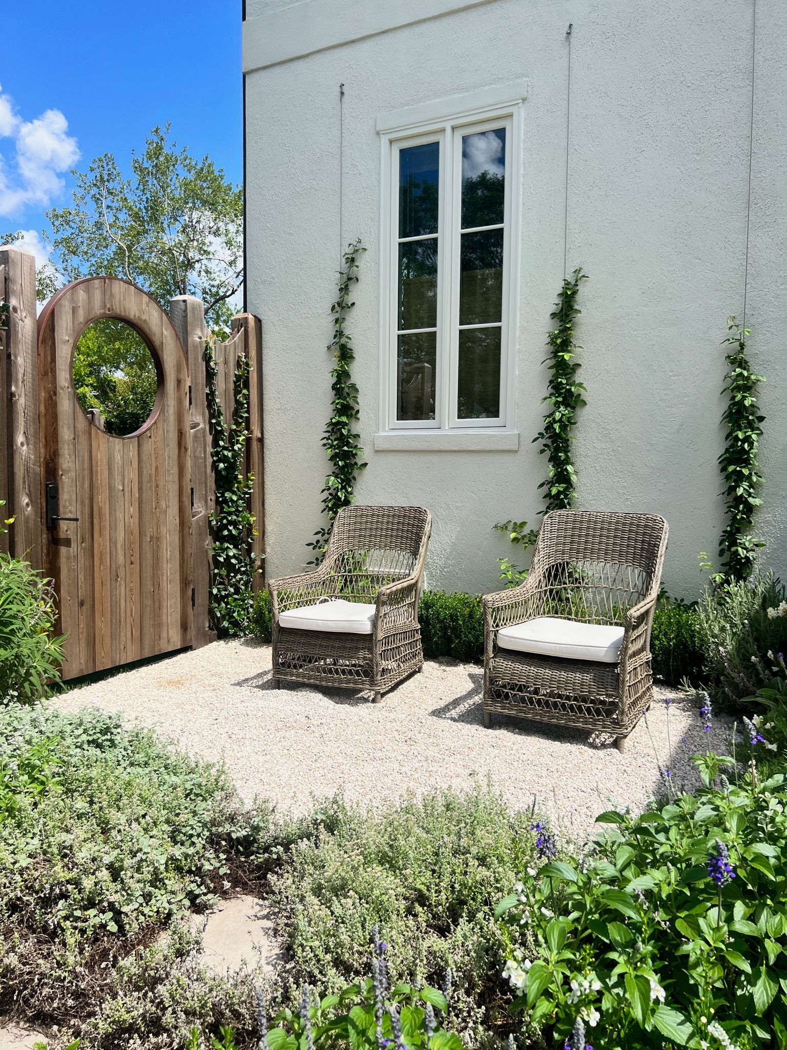 Small garden patio with two chairs, white stucco wall, window, and wooden gate with wreath