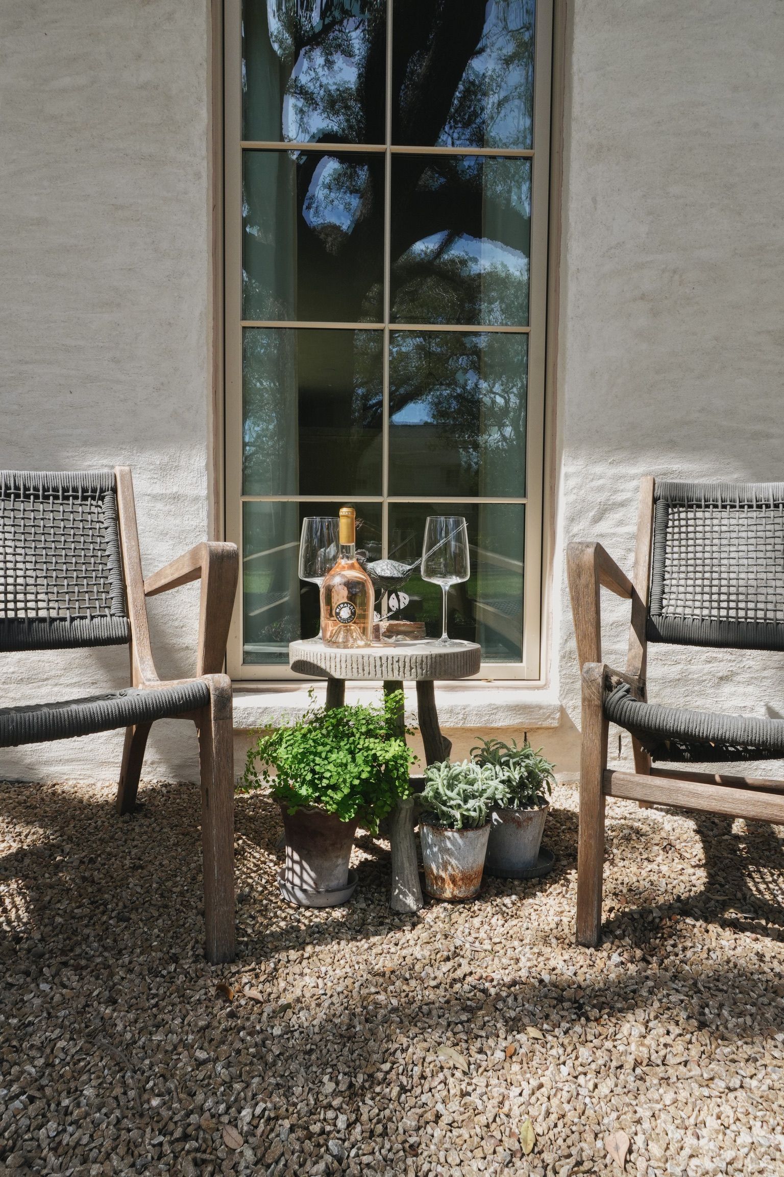 Two wicker chairs flank a small table with drinks and plants in front of a large window outside