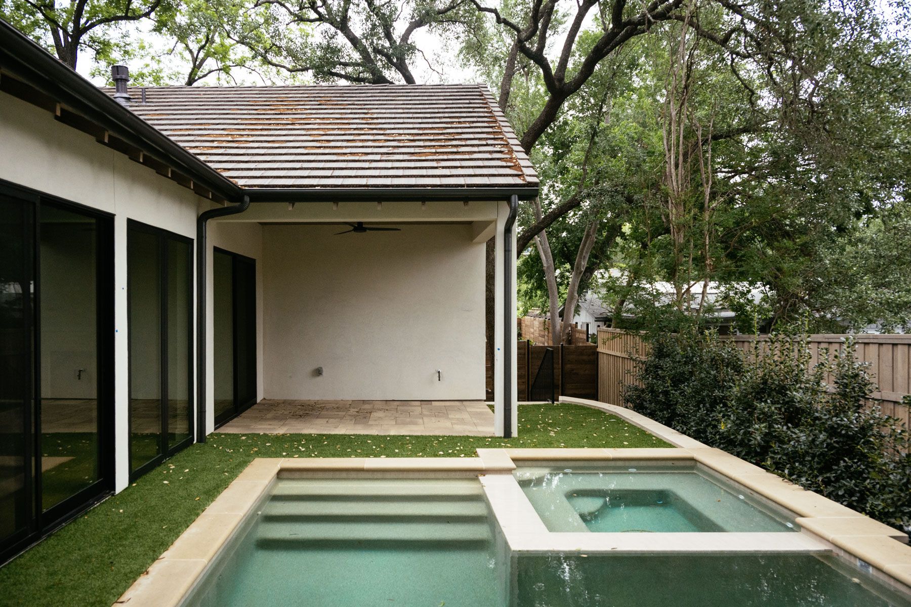 Modern backyard patio with a small rectangular pool beside a house and trees overhead
