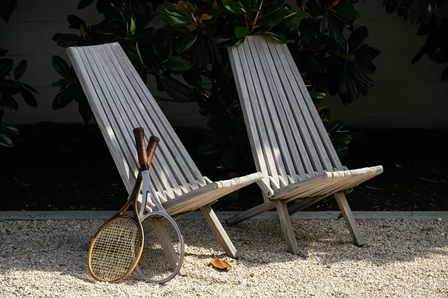 Two wooden lounge chairs on gravel with a tennis racket leaning between them outdoors