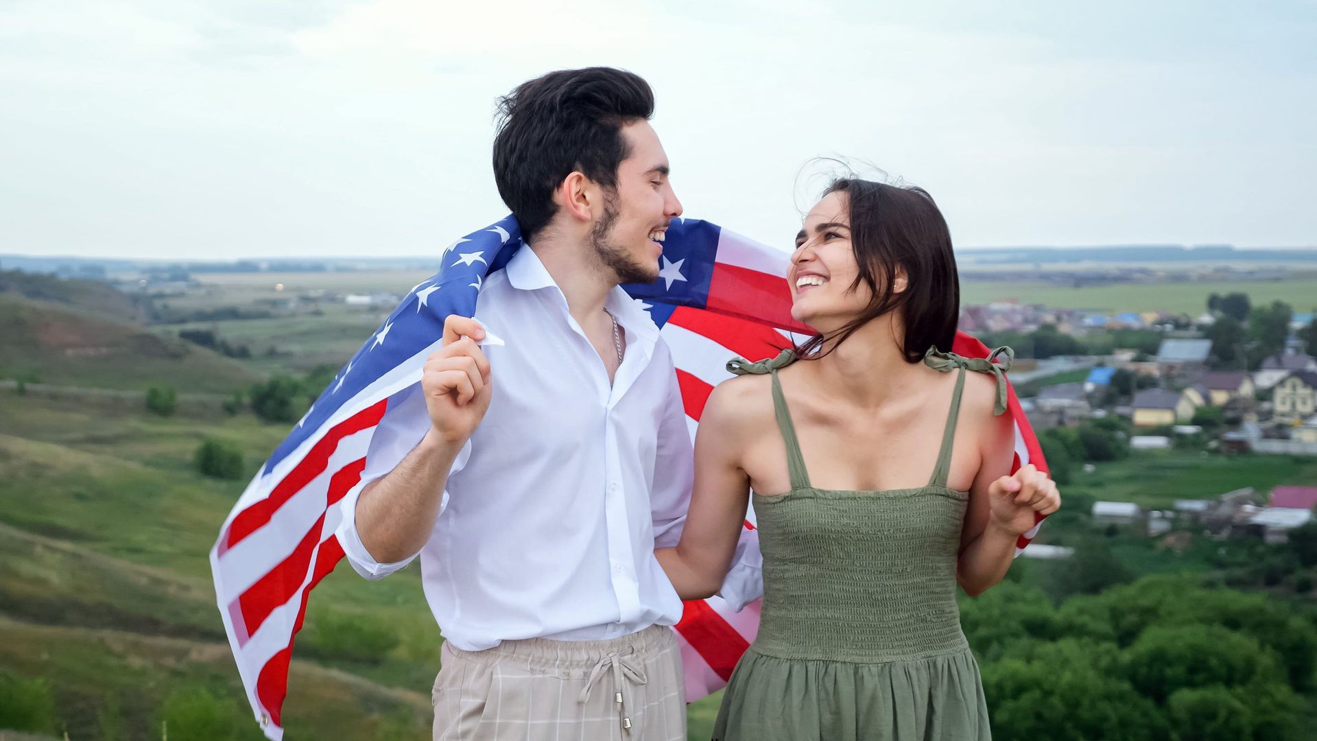 Couple outdoors with a U.S. flag, smiling and facing each other on a hill overlooking fields