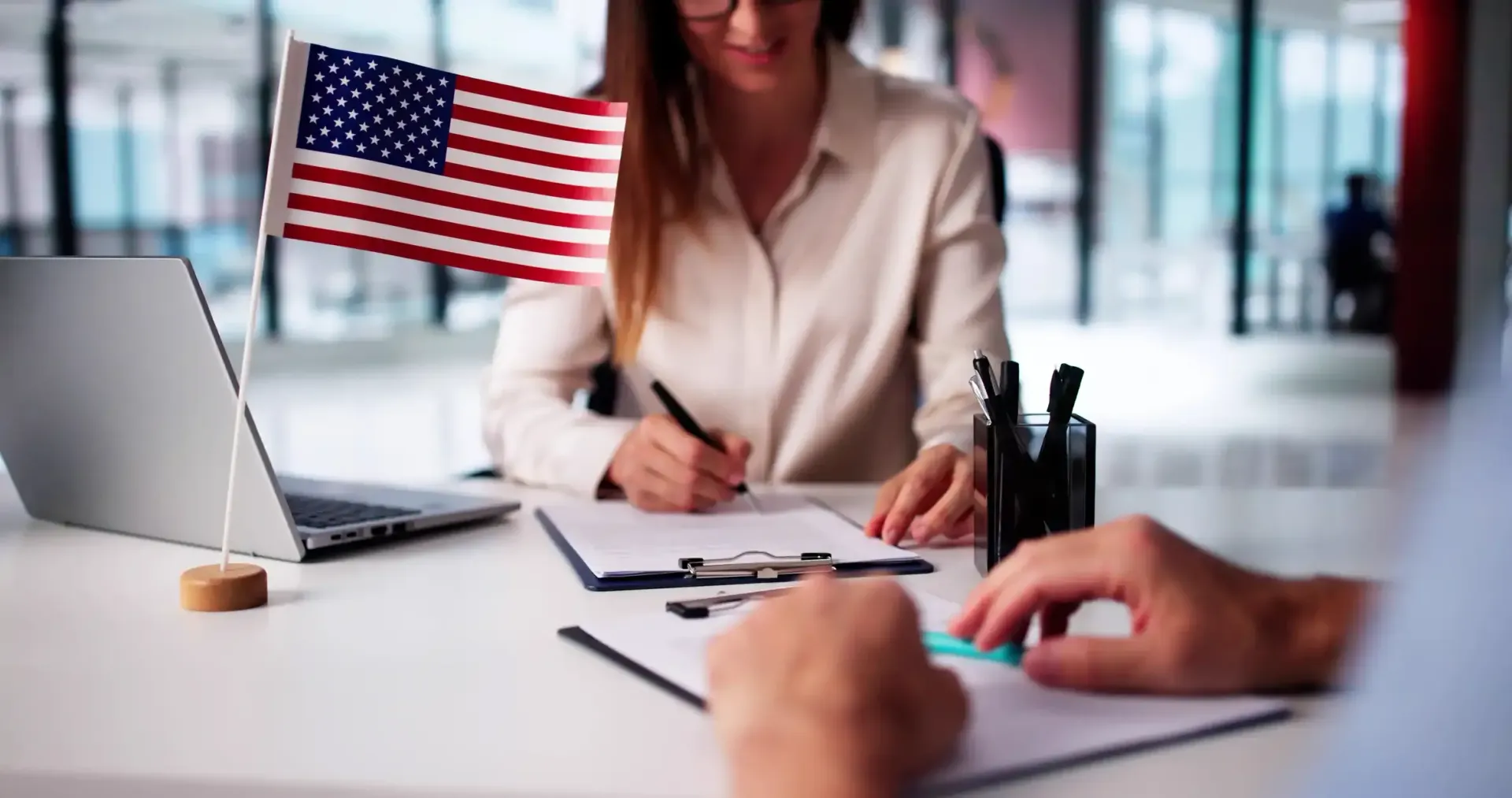 A person writes on a document at a desk with an American flag, a laptop, and a partner’s hands visible in the foreground.