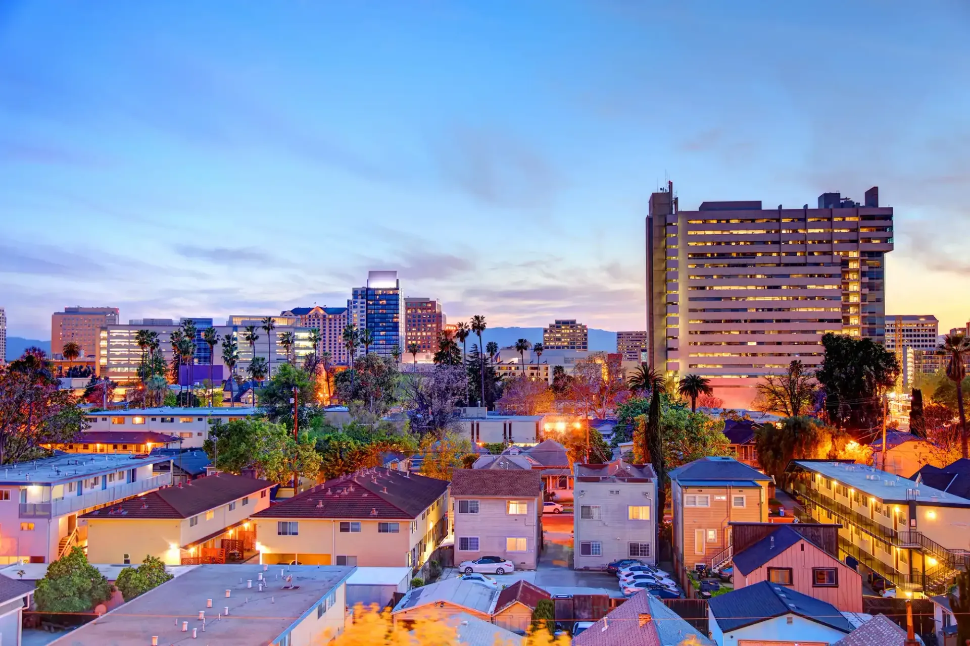 A cityscape at dusk with warm residential lights below, transitioning to taller office buildings under a blue sky.