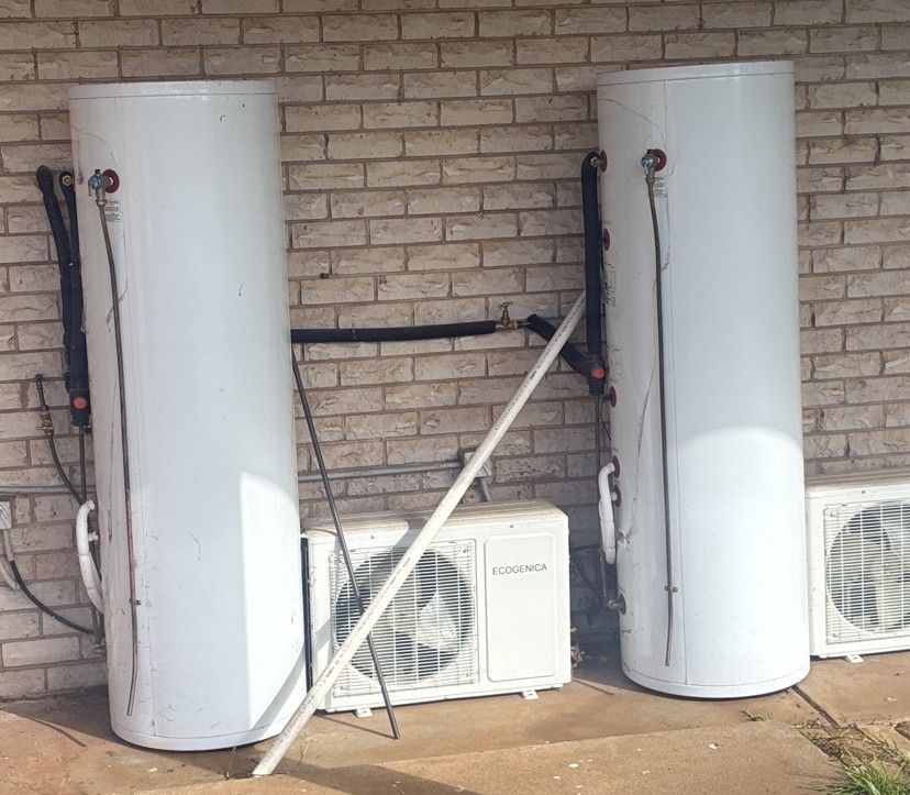 Two tall white water heaters beside outdoor air-conditioning units against a brick wall.