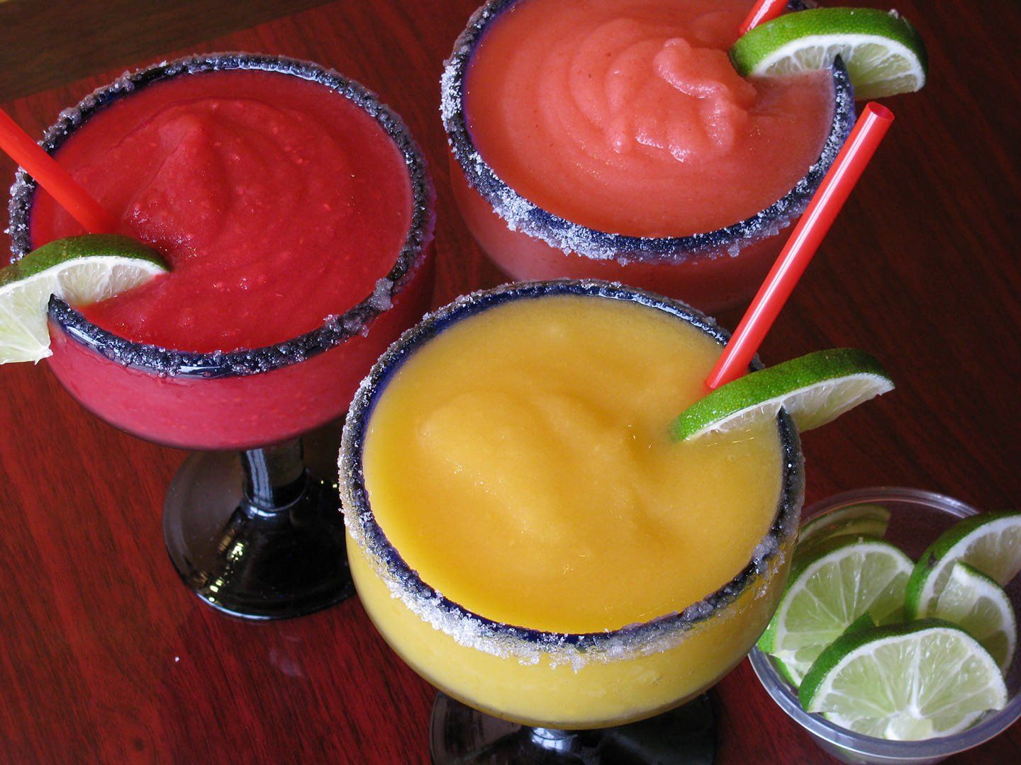 Three colorful margaritas: red, pink, and yellow. Each has a lime wedge and a red straw, salt rim.