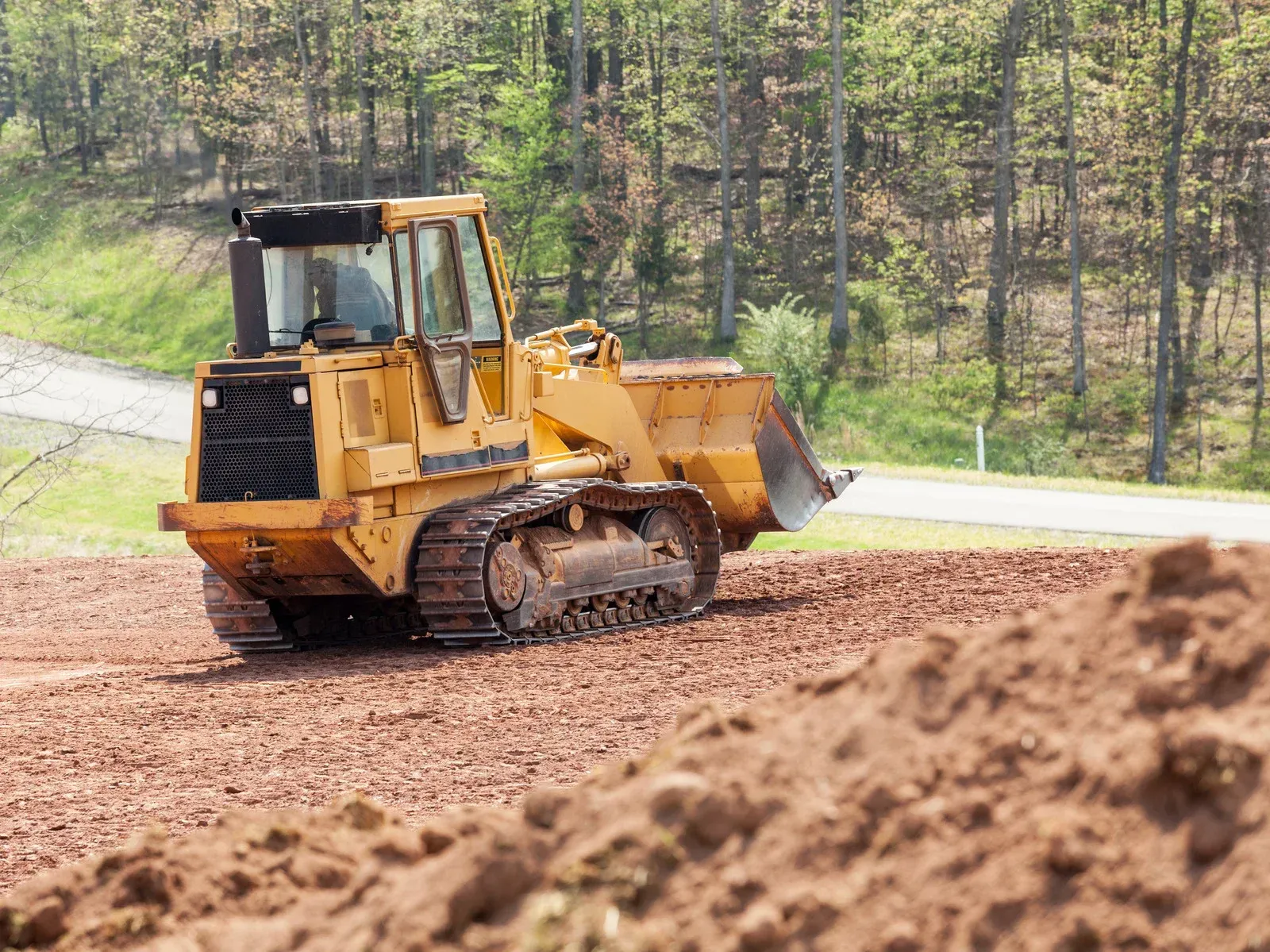 A yellow track loader sits on a patch of red dirt at a construction site with a forest in the background.