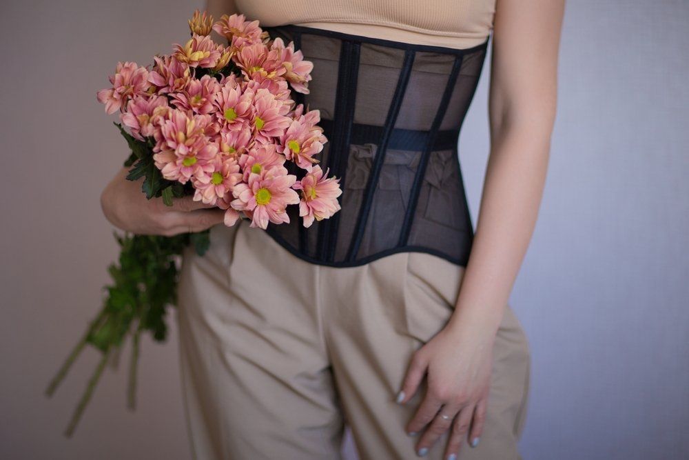 Woman in a Corset and Tan Pants Holding Pink Flowers Against a Light-coloured Wall — Shona's Lingerie in Hermit Park, QLD