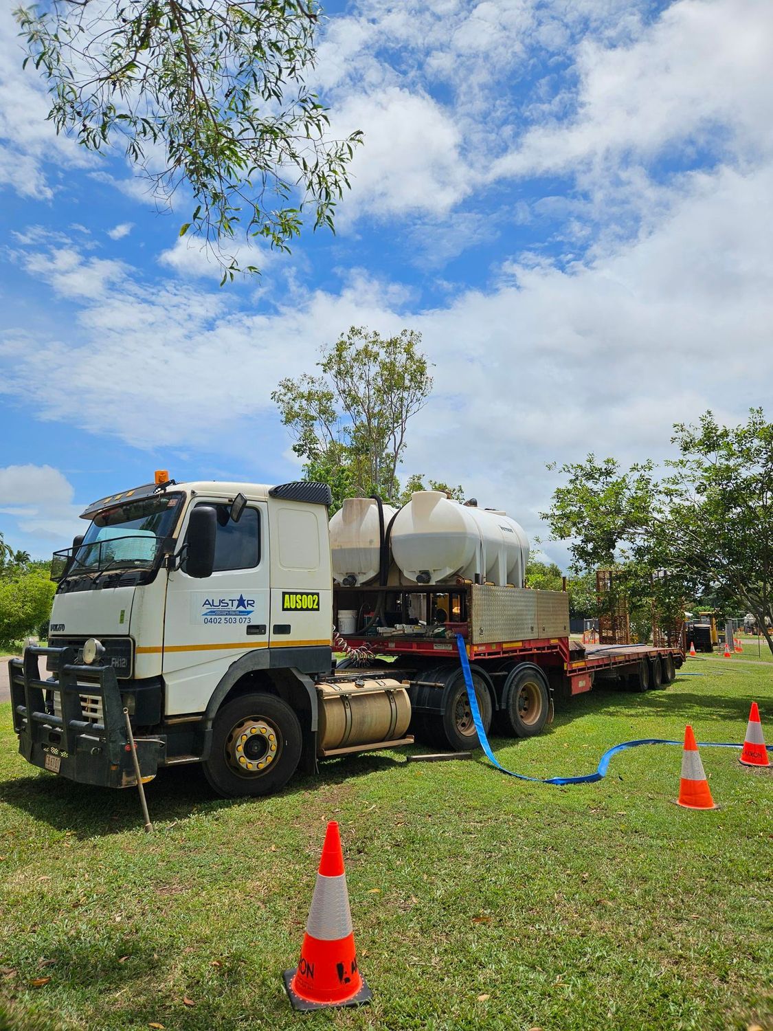 A truck carrying two large white tanks on a trailer, parked on grass with blue hoses and orange cones. — Austar Underground Services in Girraween, NT