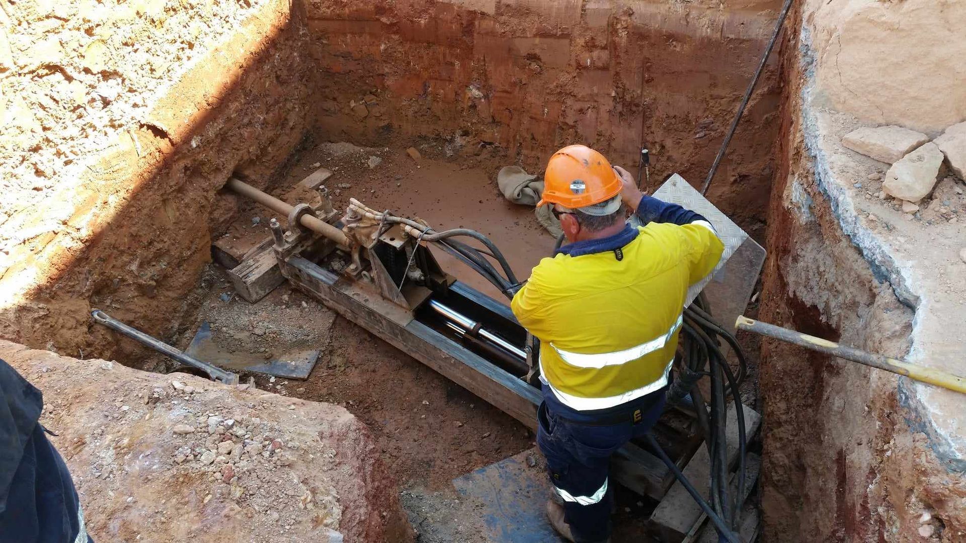 A worker in a pit using a drill to drill horizontally into the pit wall.
