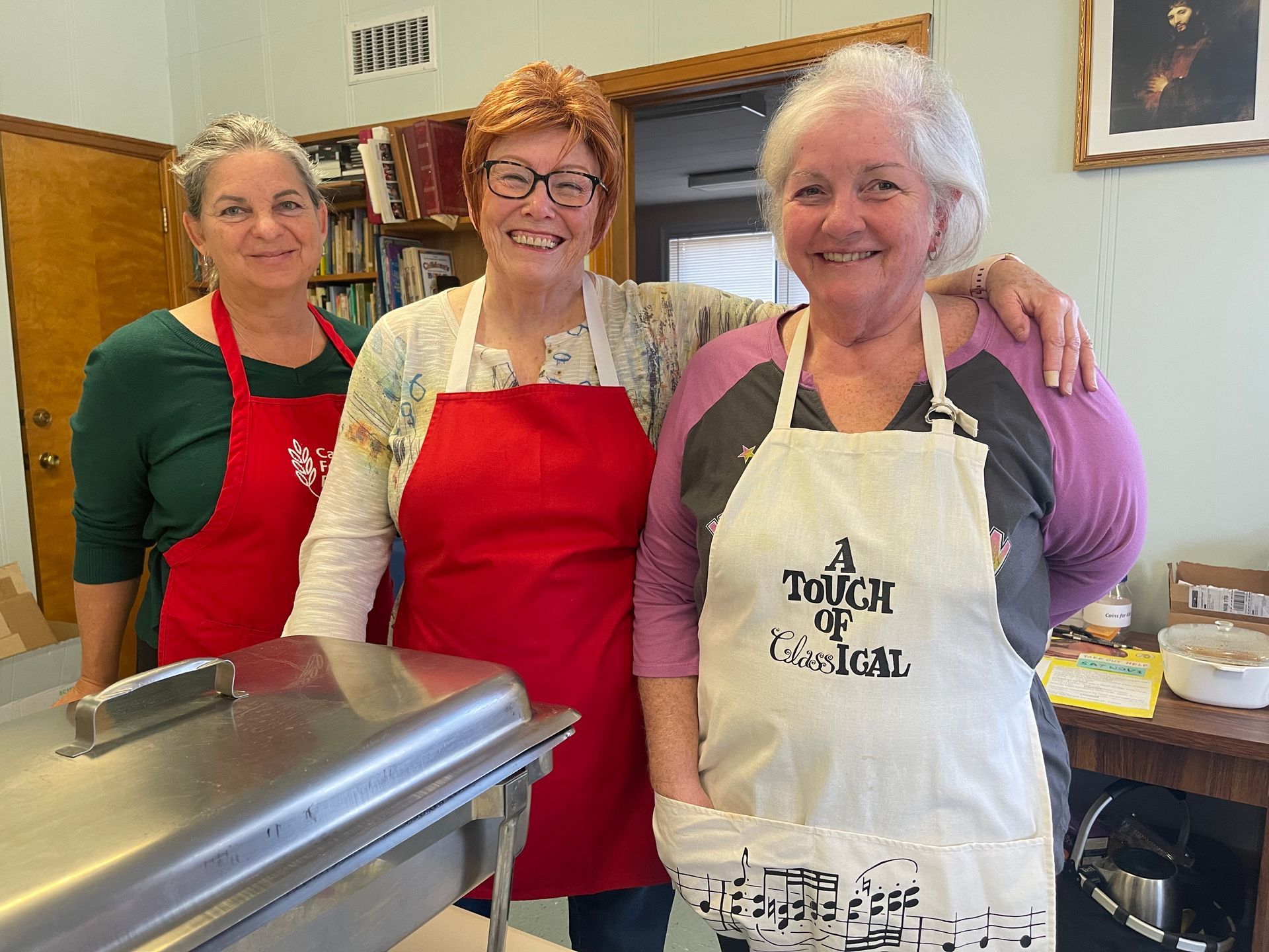 Three women smiling in a kitchen, wearing aprons and standing behind a counter.