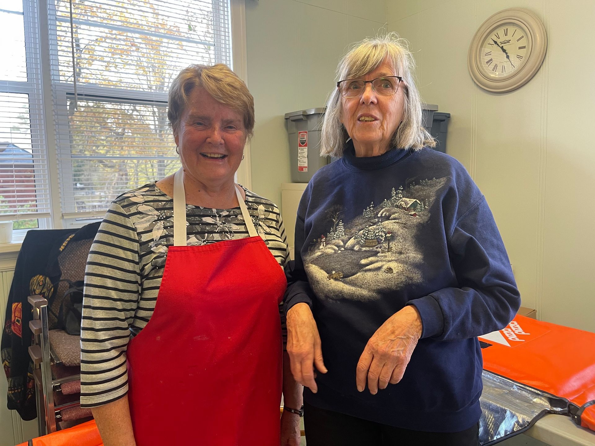 Two women smiling in a kitchen, one in a red apron and one in a navy sweatshirt.