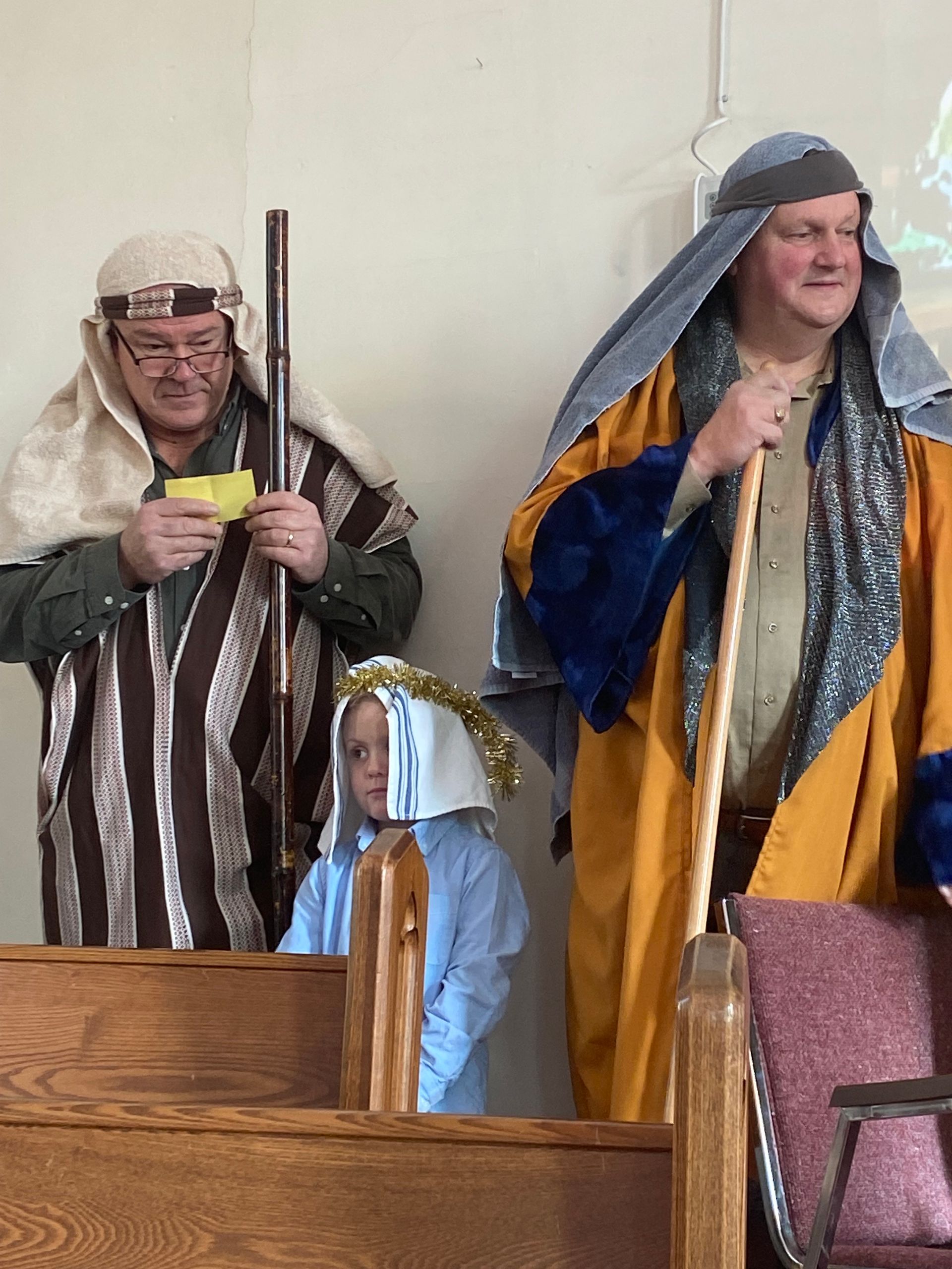 People in medieval costumes standing indoors near wooden pews, one holding a staff, one in a blue robe