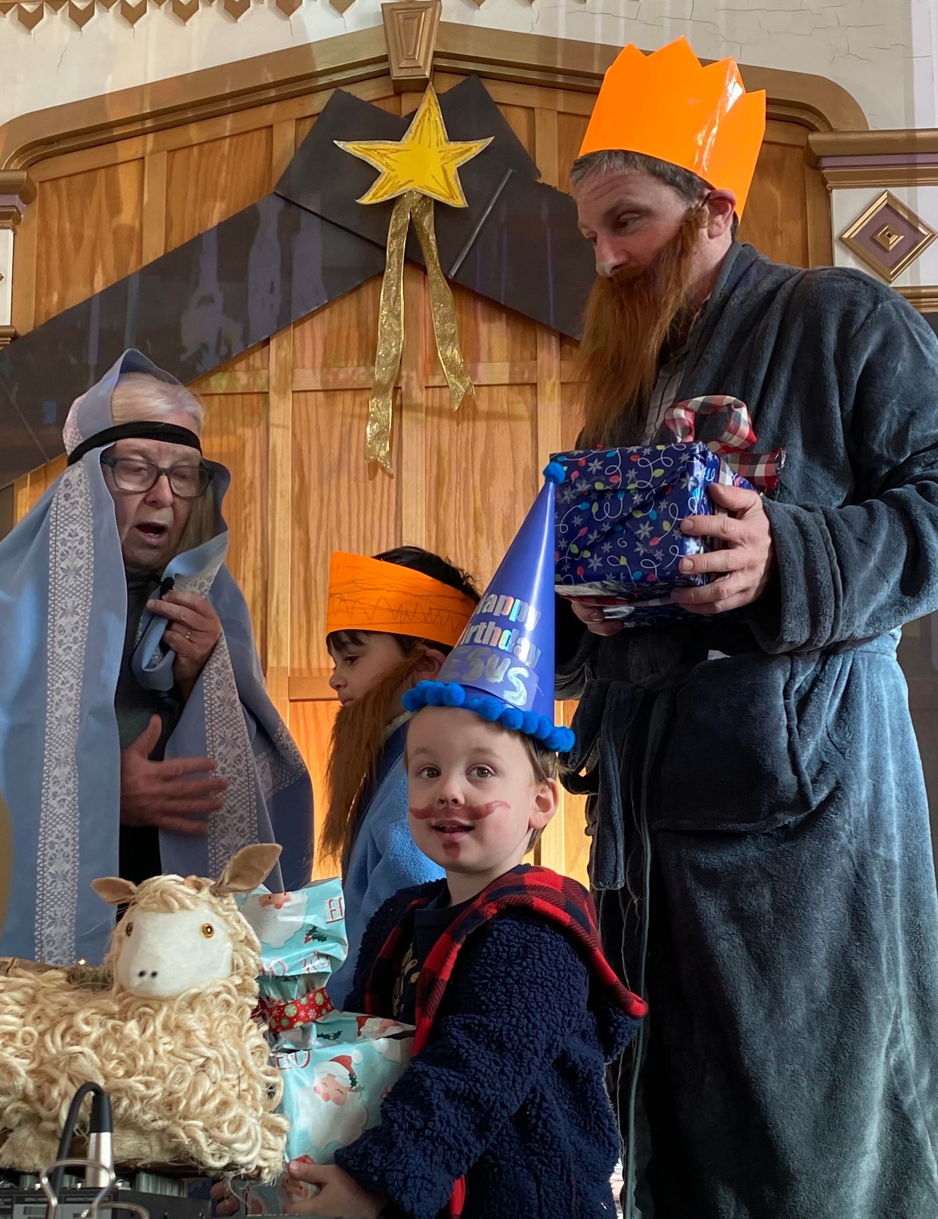 People in festive costumes around a child in a blue party hat holding a wrapped gift indoors