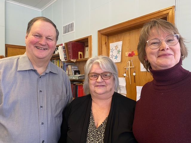 Three smiling people standing indoors by a wooden door and shelf.