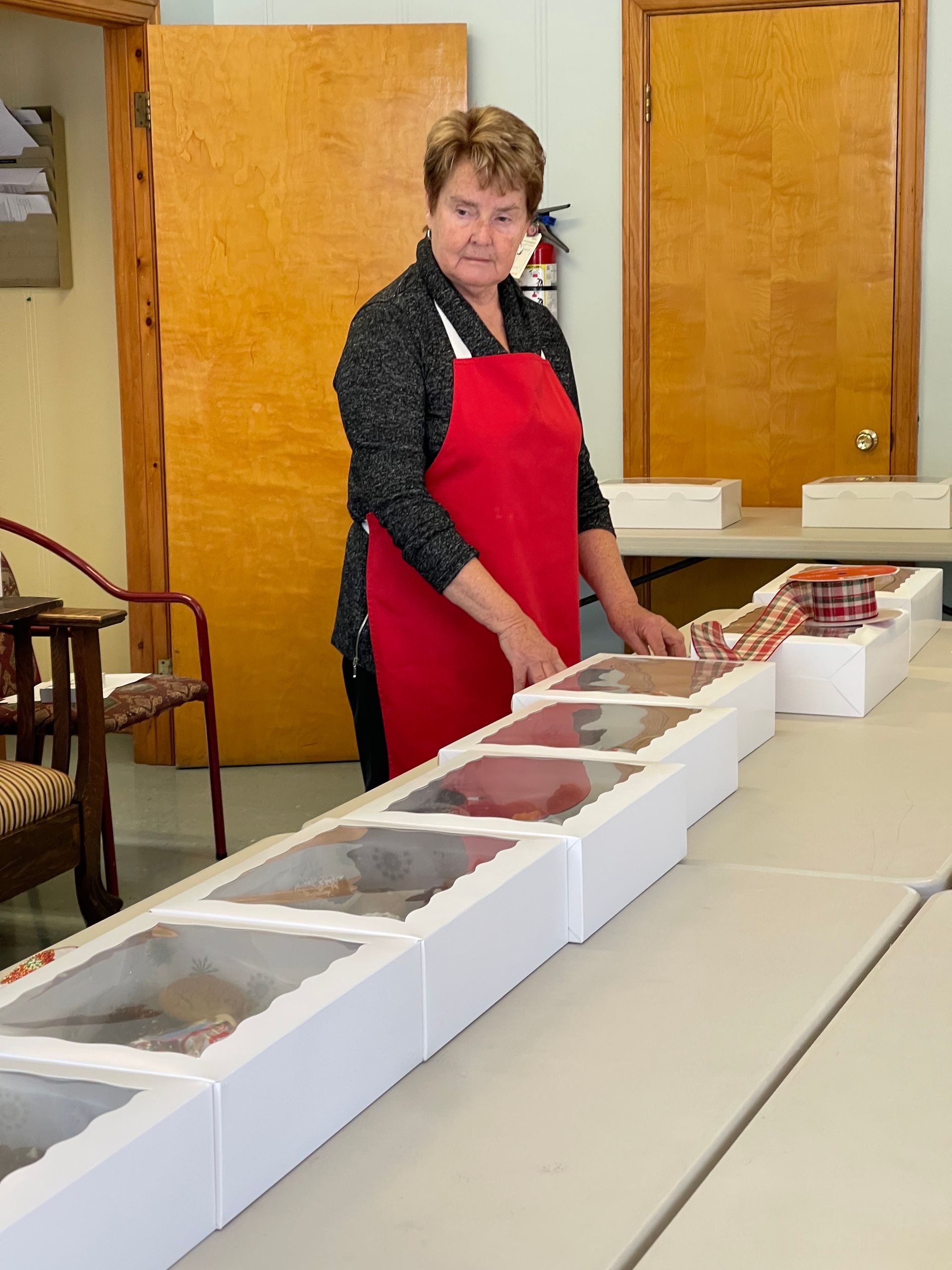 Woman in a red apron arranging food trays on a table in a room with wooden cabinets
