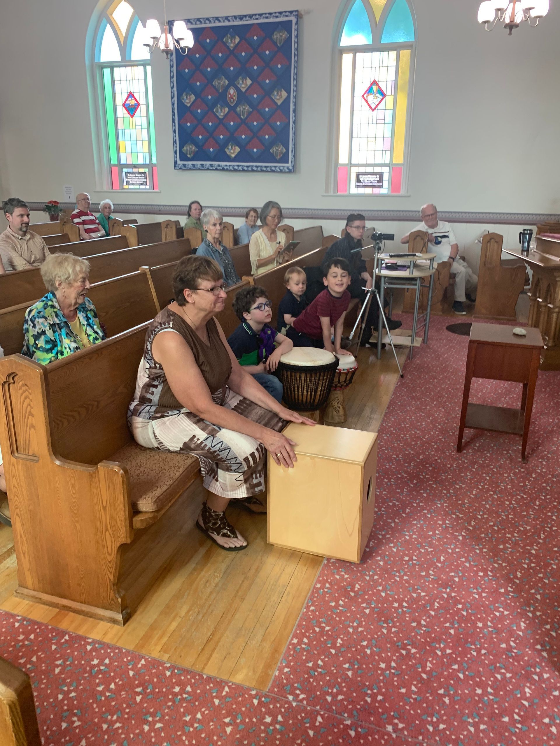 People seated in a church pew facing an altar, with stained-glass windows and a red carpet.