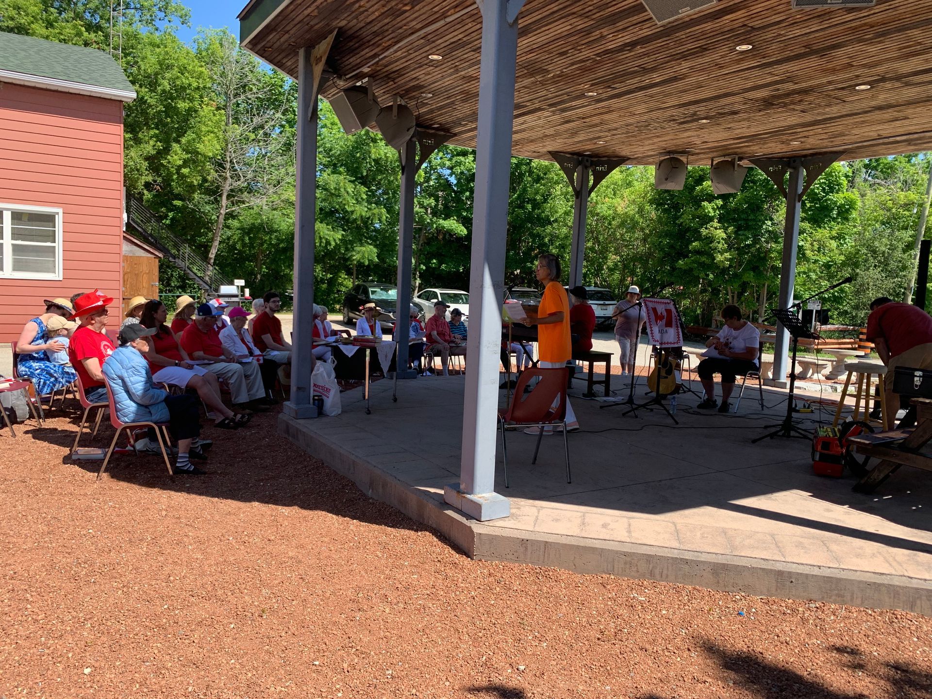 People seated at picnic tables under a pavilion beside a red brick building on a sunny day