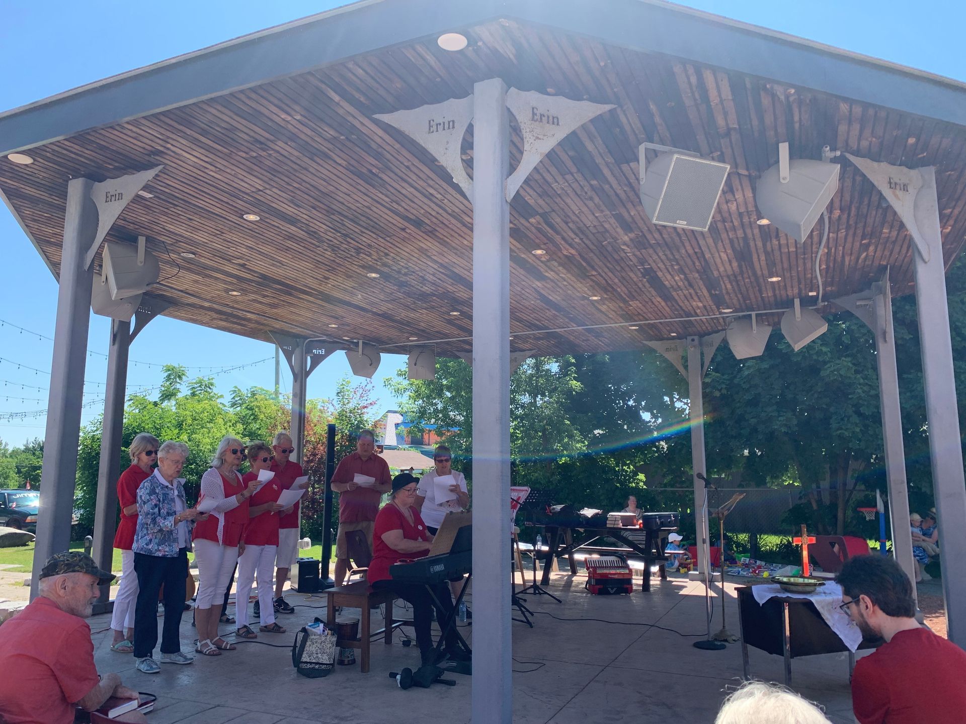 Outdoor stage with musicians and microphones under a wooden pavilion in bright sunlight