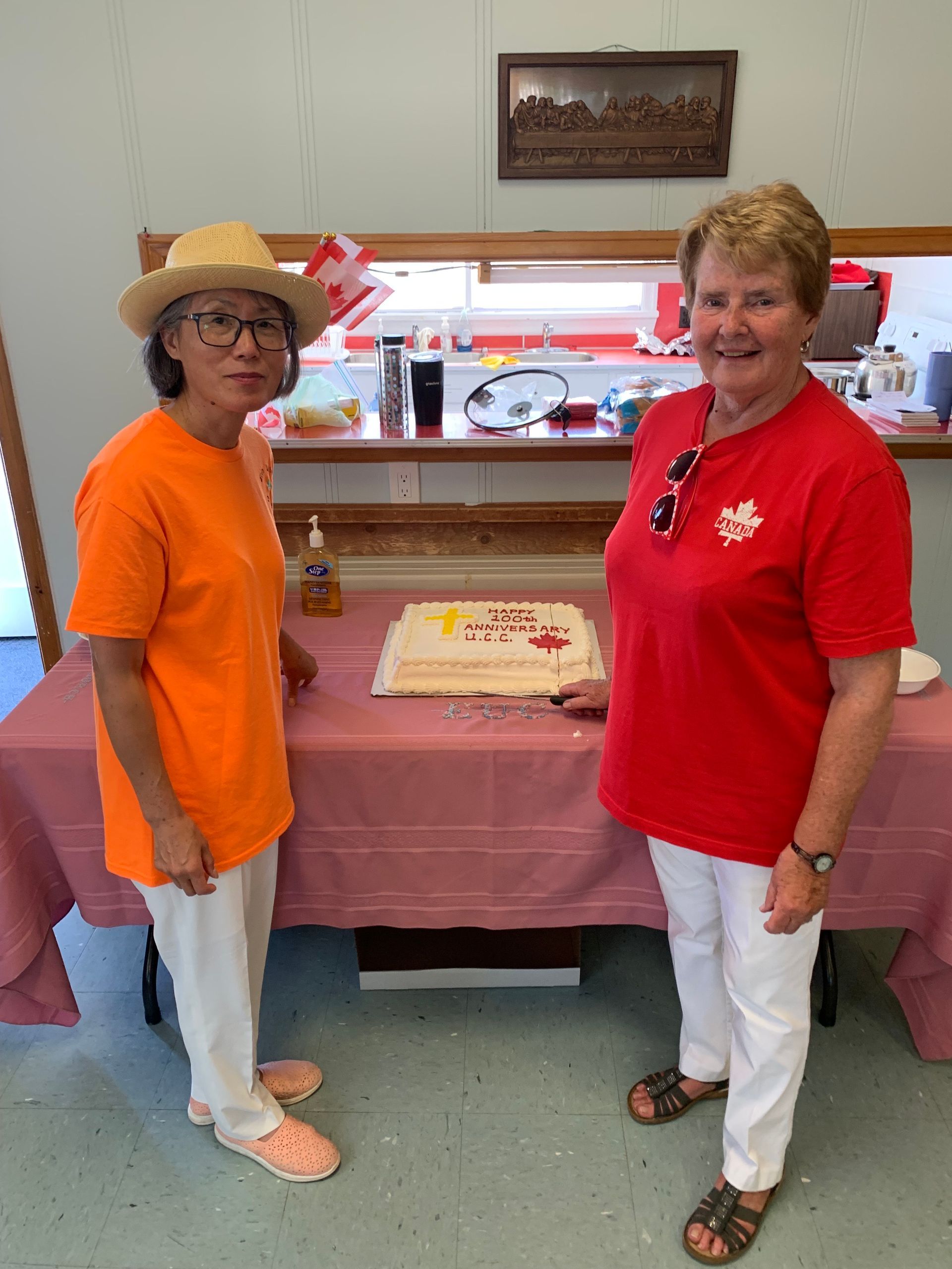 Two women standing beside a decorated cake on a table in a room, smiling at the camera.