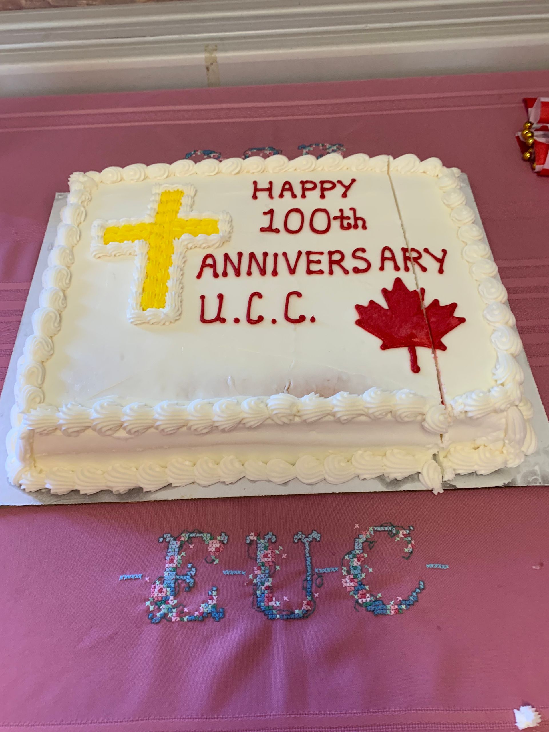 White cake with yellow cross, red maple leaf, and “Happy 100th Anniversary U.C.C.” on a pink tablecloth.