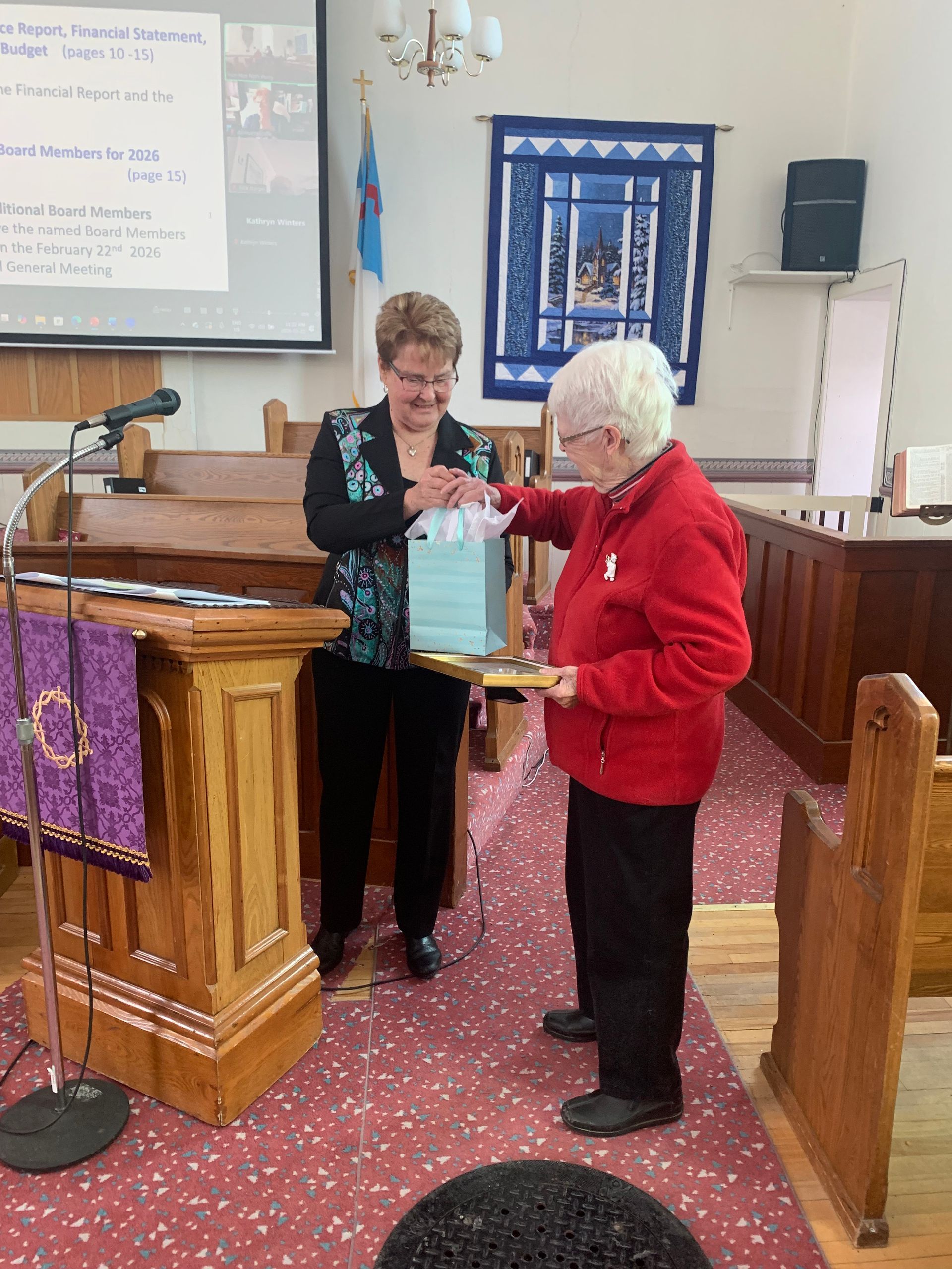 Two women exchange an item in a church sanctuary beside a wooden pulpit and pews.