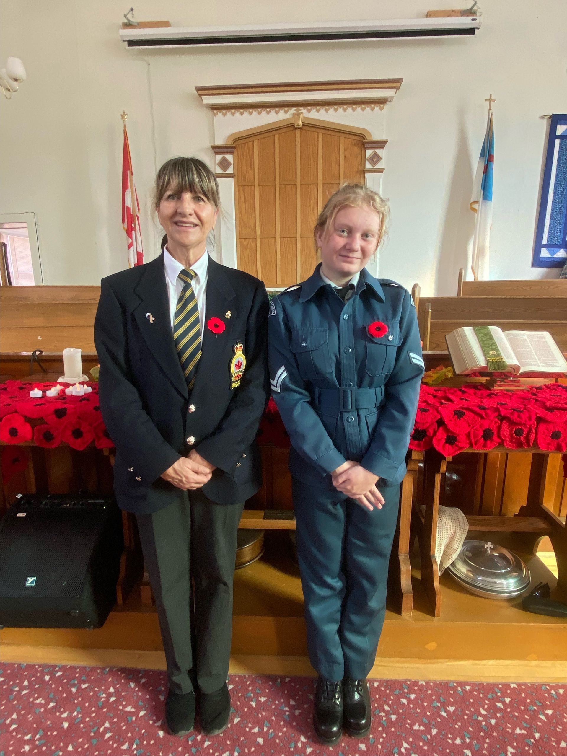 Two people standing indoors in dark uniforms, smiling in front of a wooden lectern and flags.