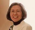 Smiling person with glasses and shoulder-length hair in a cream sweater against a light background