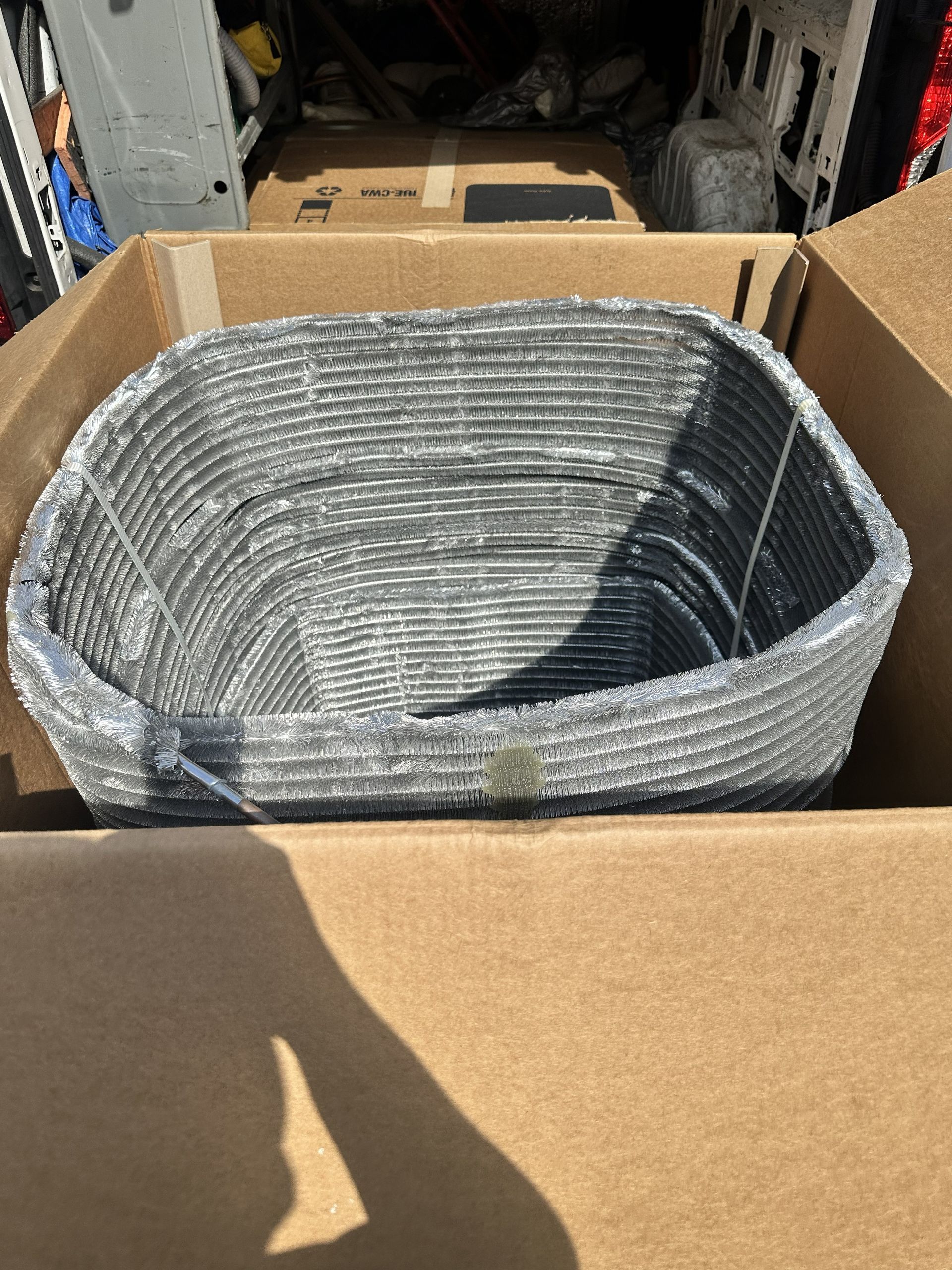 Gray woven basket in a cardboard box, inside a vehicle trunk.