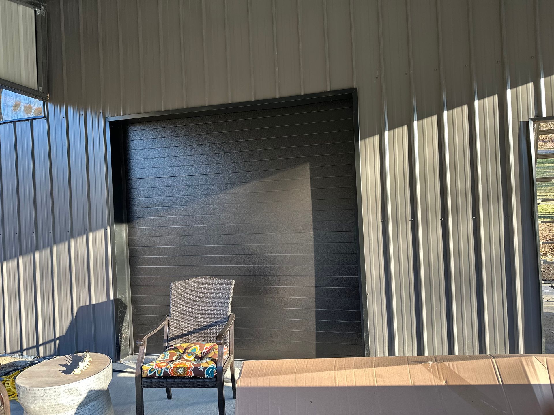 Outdoor patio with a chair, small table, and dark garage door on a corrugated metal wall.